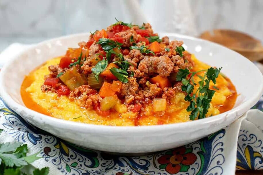 A bowl of polenta topped with a hearty Skillet Osso Buco-style meat and vegetable sauce, garnished with fresh parsley, sits on a patterned napkin with herbs on the side.