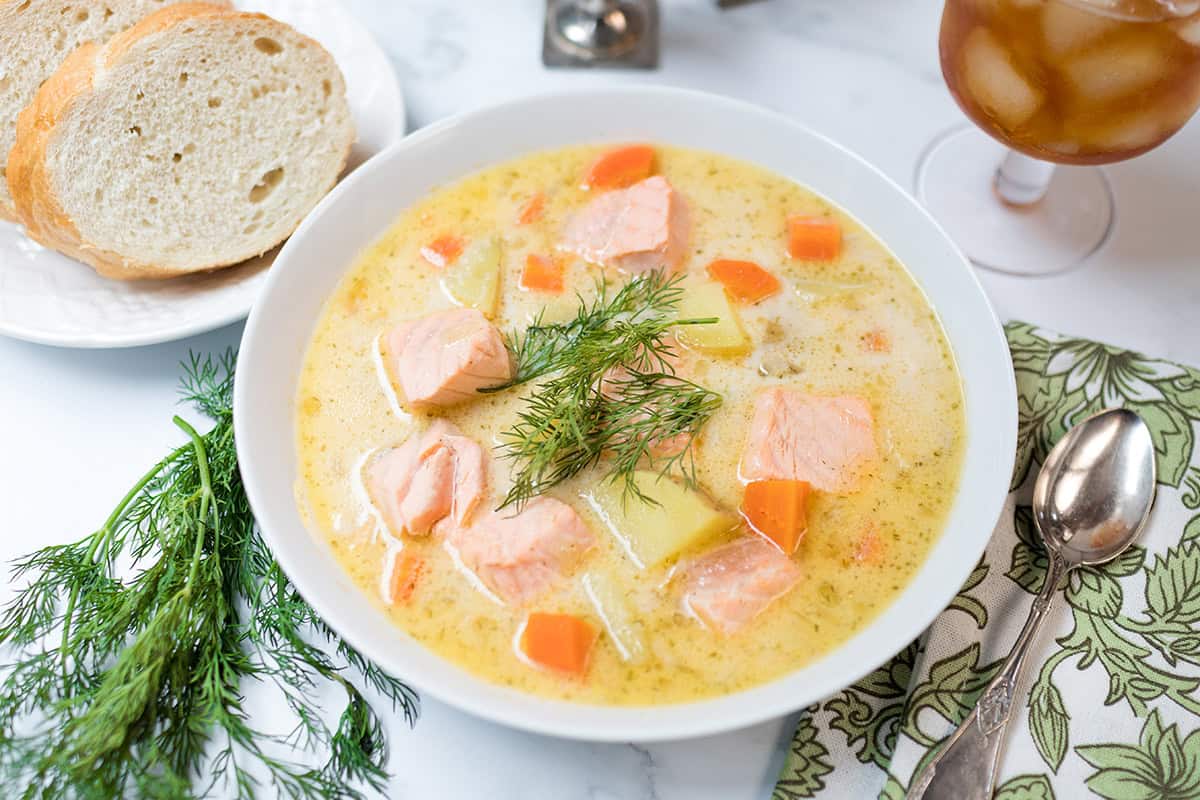 A bowl of creamy Finnish Salmon Soup with vegetables, garnished with dill, served alongside sliced bread and iced tea on a marble table.