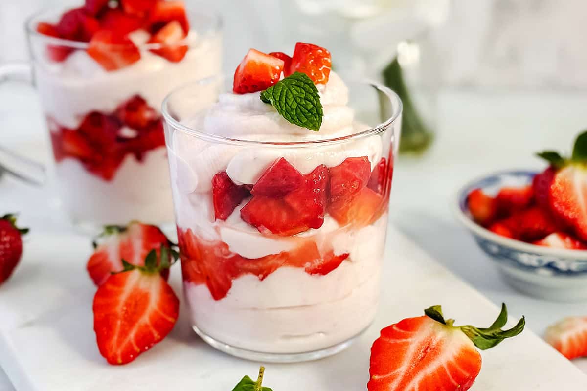 A glass cup filled with layers of honey-sweetened whipped cream and sliced strawberries, creating a delightful strawberry fool, topped with a mint leaf and surrounded by additional strawberries.