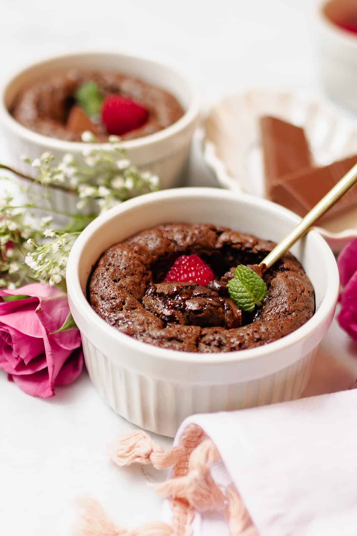 A chocolate lava cake in a white ramekin, made with a mini chocolate cake recipe, is garnished with a raspberry and mint leaf. A spoon is inserted, while pink roses, chocolate pieces, and a pink napkin sit nearby.