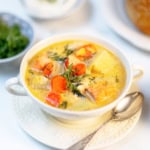 A bowl of Finnish Salmon Soup (Lohikeitto) with chunks of salmon, potatoes, carrots, and fresh dill, served on a white saucer with a spoon, with herbs and bread in the background.