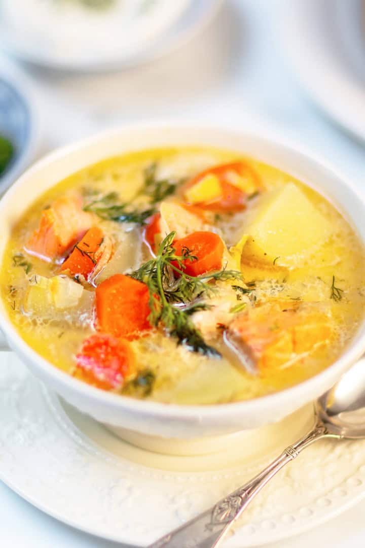 A bowl of Finnish Salmon Soup (Lohikeitto) with chunks of salmon, potatoes, carrots, and fresh dill, served on a white saucer with a spoon, with herbs and bread in the background.