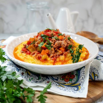 A bowl of creamy polenta topped with a chunky Skillet Osso Buco meat and vegetable sauce, garnished with fresh parsley, on a decorative napkin.