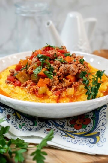 A bowl of creamy polenta topped with a chunky Skillet Osso Buco meat and vegetable sauce, garnished with fresh parsley, on a decorative napkin.