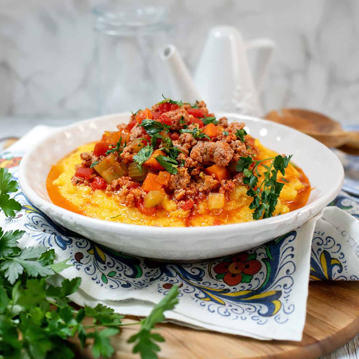 A bowl of creamy polenta topped with a chunky Skillet Osso Buco meat and vegetable sauce, garnished with fresh parsley, on a decorative napkin.