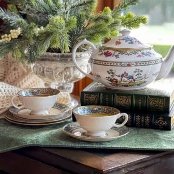 A floral teapot and two teacups sit on stacked books beside a vase of greenery, all arranged on a green tablecloth near a window-a serene still life speaking the language of love.
