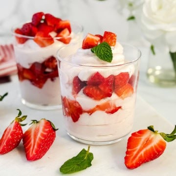 Two clear glasses filled with layers of honey-sweetened whipped cream, yogurt, and chopped strawberries, garnished with a mint leaf, with extra strawberries and mint on the table-a delicious twist on classic strawberry fool.