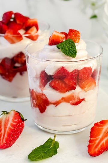 Two clear glasses filled with layers of honey-sweetened whipped cream, yogurt, and chopped strawberries, garnished with a mint leaf, with extra strawberries and mint on the table—a delicious twist on classic strawberry fool.