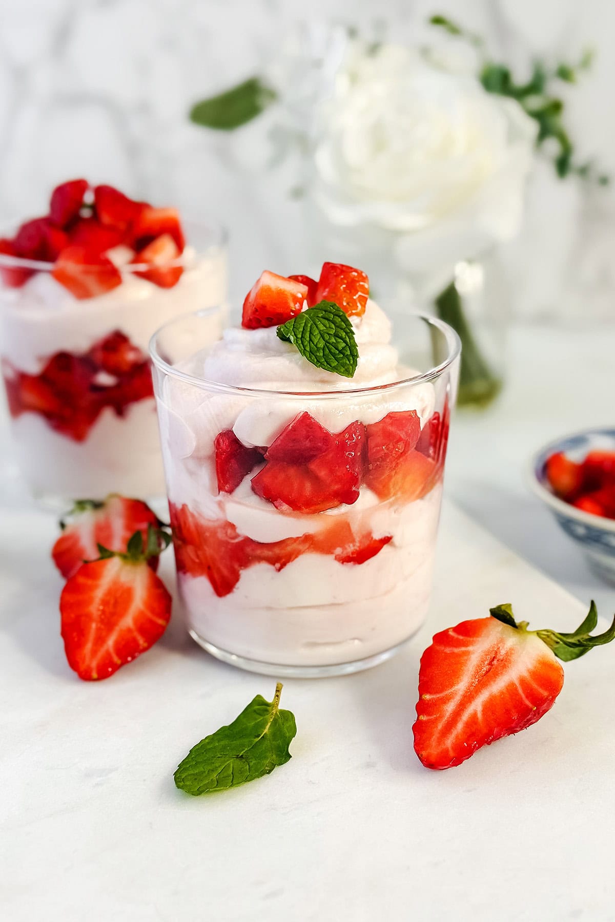 Two glass cups filled with layers of honey sweetened whipped cream and chopped strawberries, inspired by a classic strawberry fool, garnished with mint. Whole strawberries and a mint leaf rest on the table, with a white rose in the background.