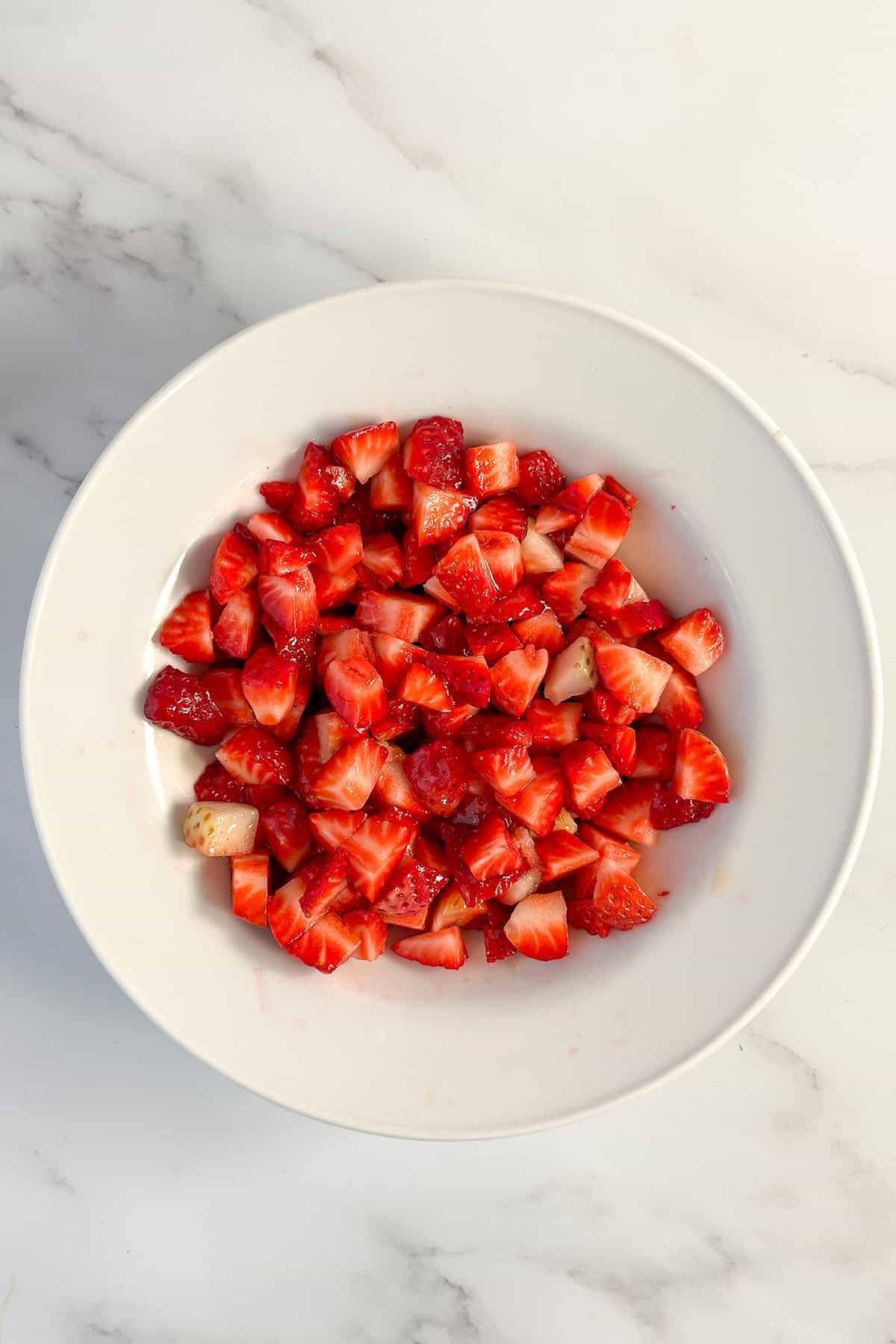 A white bowl filled with chopped fresh strawberries sits on a white marble surface, perfect for a light strawberry dessert or as a topping for your favorite yogurt dessert.