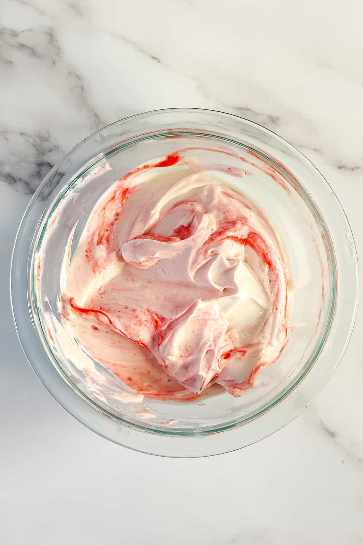 A glass bowl filled with swirled pink and white honey-sweetened strawberry fool sits on a white marble surface.