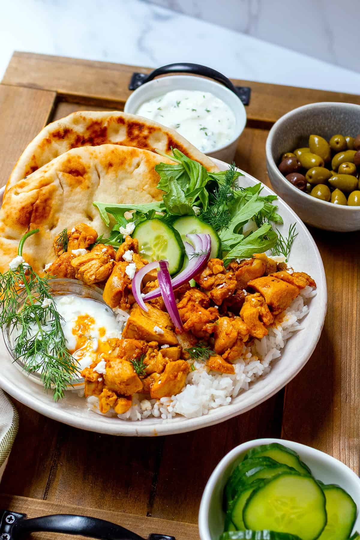 A plate with grilled Chicken Shawarma over rice, naan bread, fresh greens, cucumber, red onion, a dollop of yogurt sauce, with olives and cucumber slices on the side.