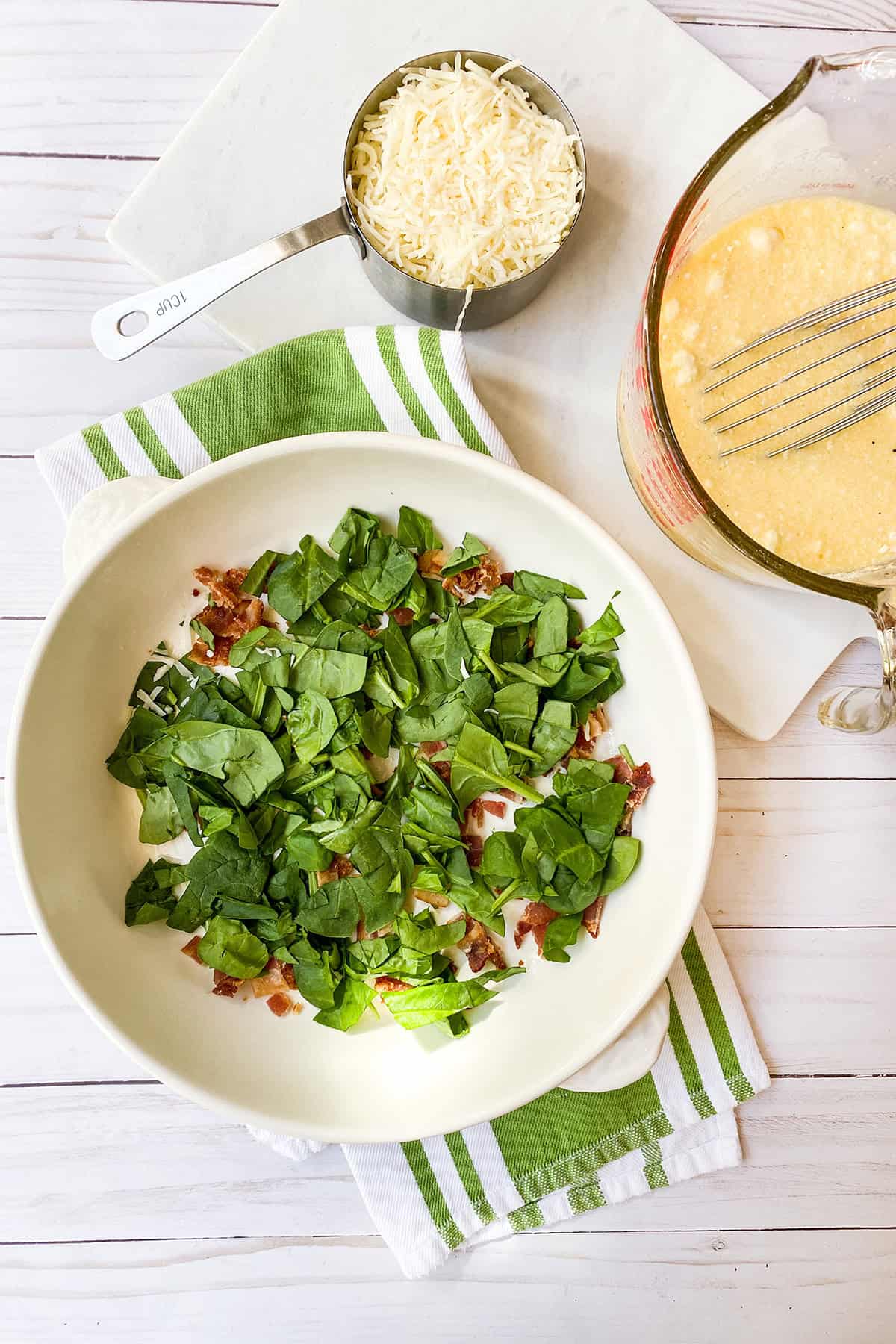 A white dish with chopped spinach and bacon, perfect for quick and easy recipes, sits next to a cup of shredded cheese and a measuring cup with beaten eggs on a striped green and white towel.