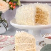 A slice of coconut cake is served on a plate in front of a whole coconut cake on a stand, with flowers and dishes in the background. Text reads "Coconut Cake" and "Get the Recipe.