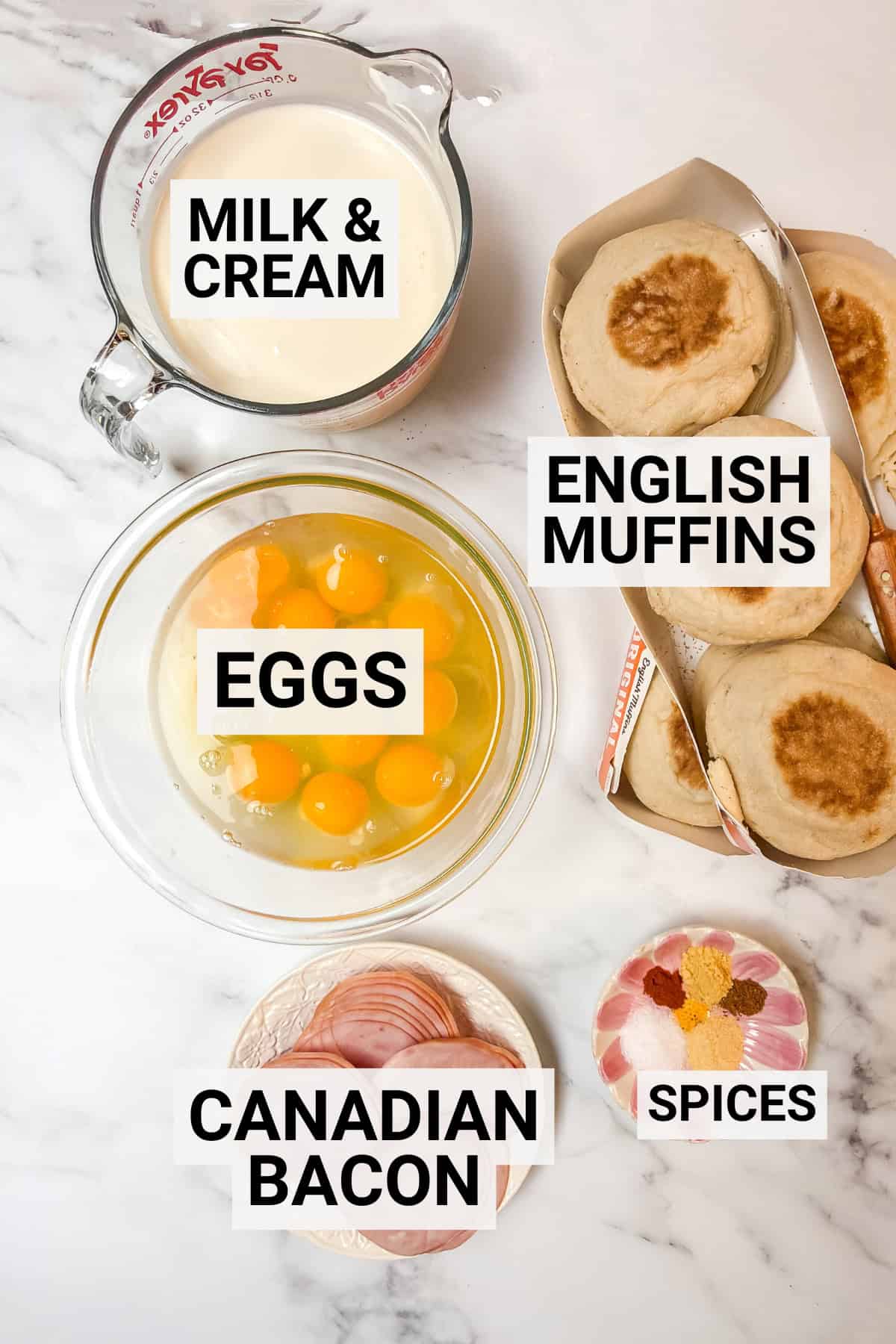 Eggs benedict casserole ingredients including english muffins, eggs, milk, cream, Canadian bacon, and spices on marble surface.