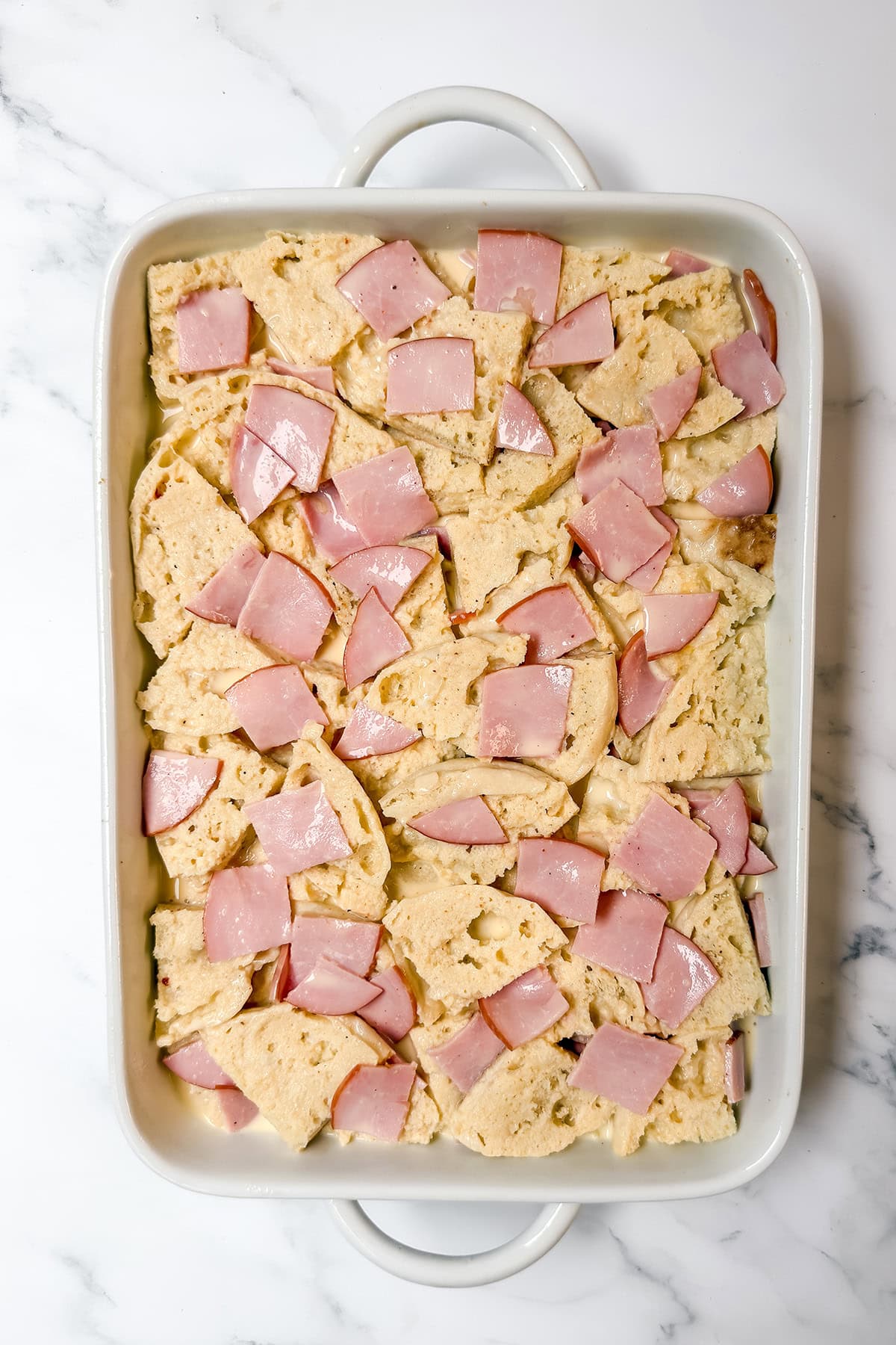 Pouring egg custard over english muffins and ham in casserole dish.
