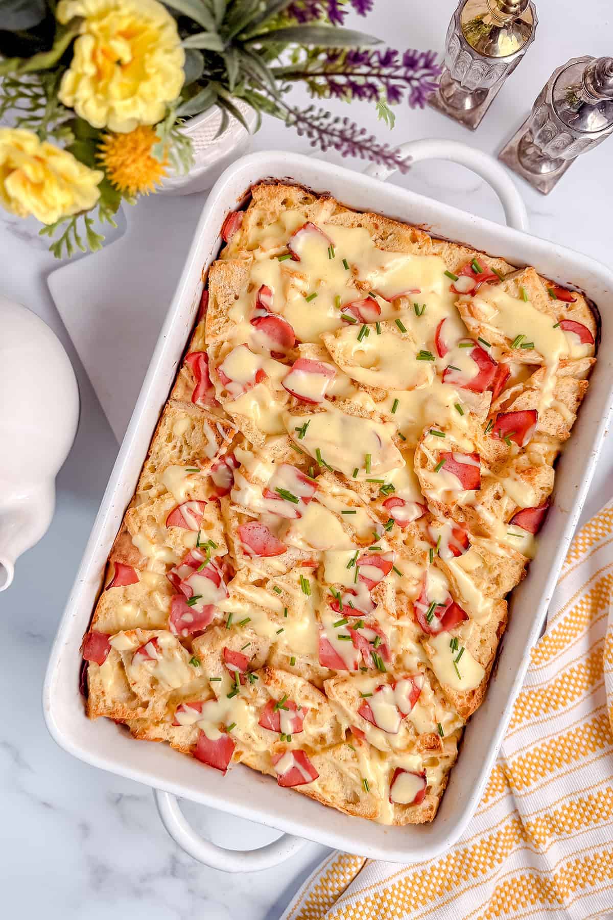 Baked eggs benedict casserole topped with hollandaise sauce and chives in white baking dish.