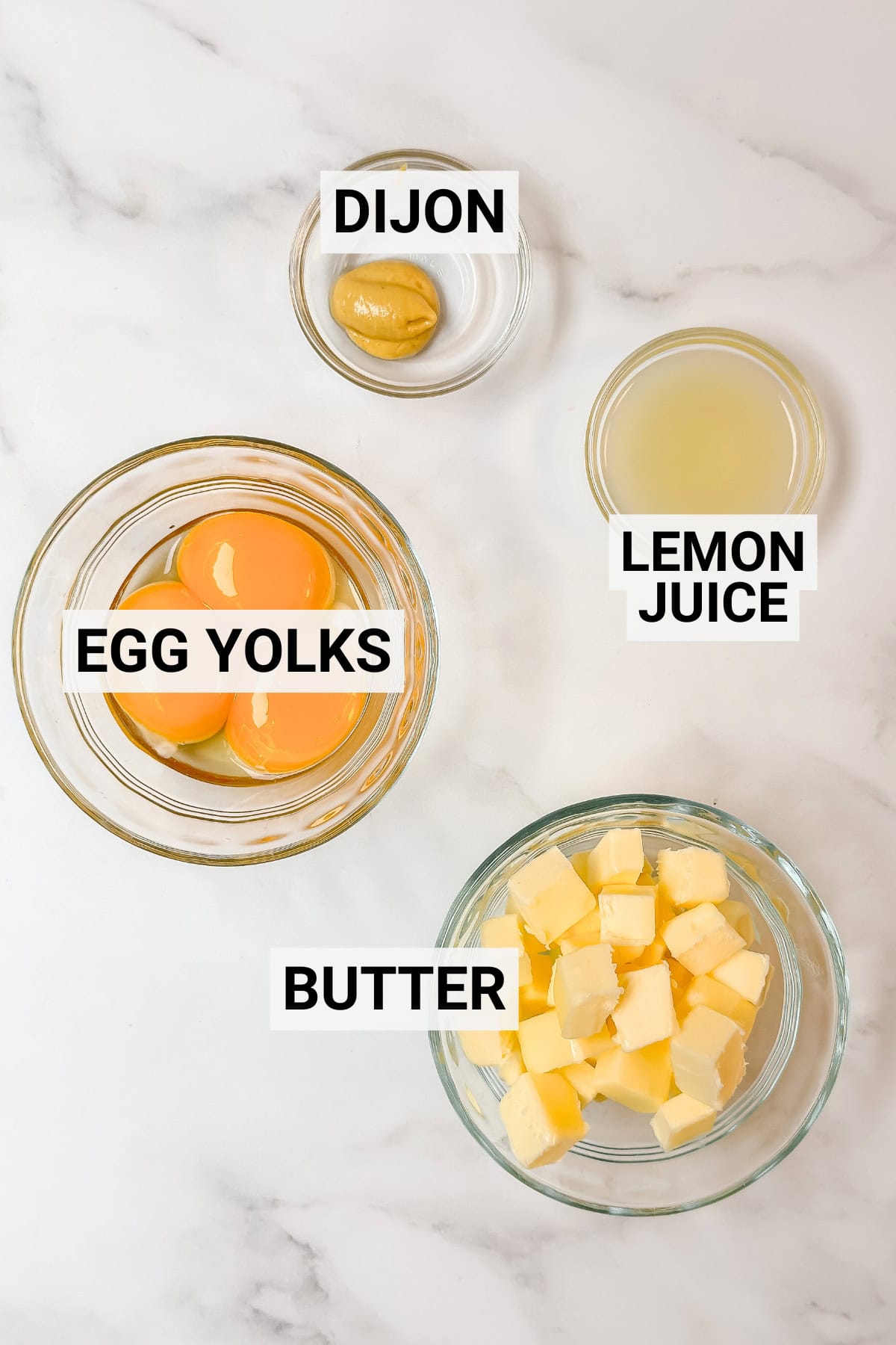 Four glass bowls on a marble surface containing labeled ingredients for Stovetop Hollandaise Sauce: egg yolks, cubes of butter, dijon mustard, and lemon juice.