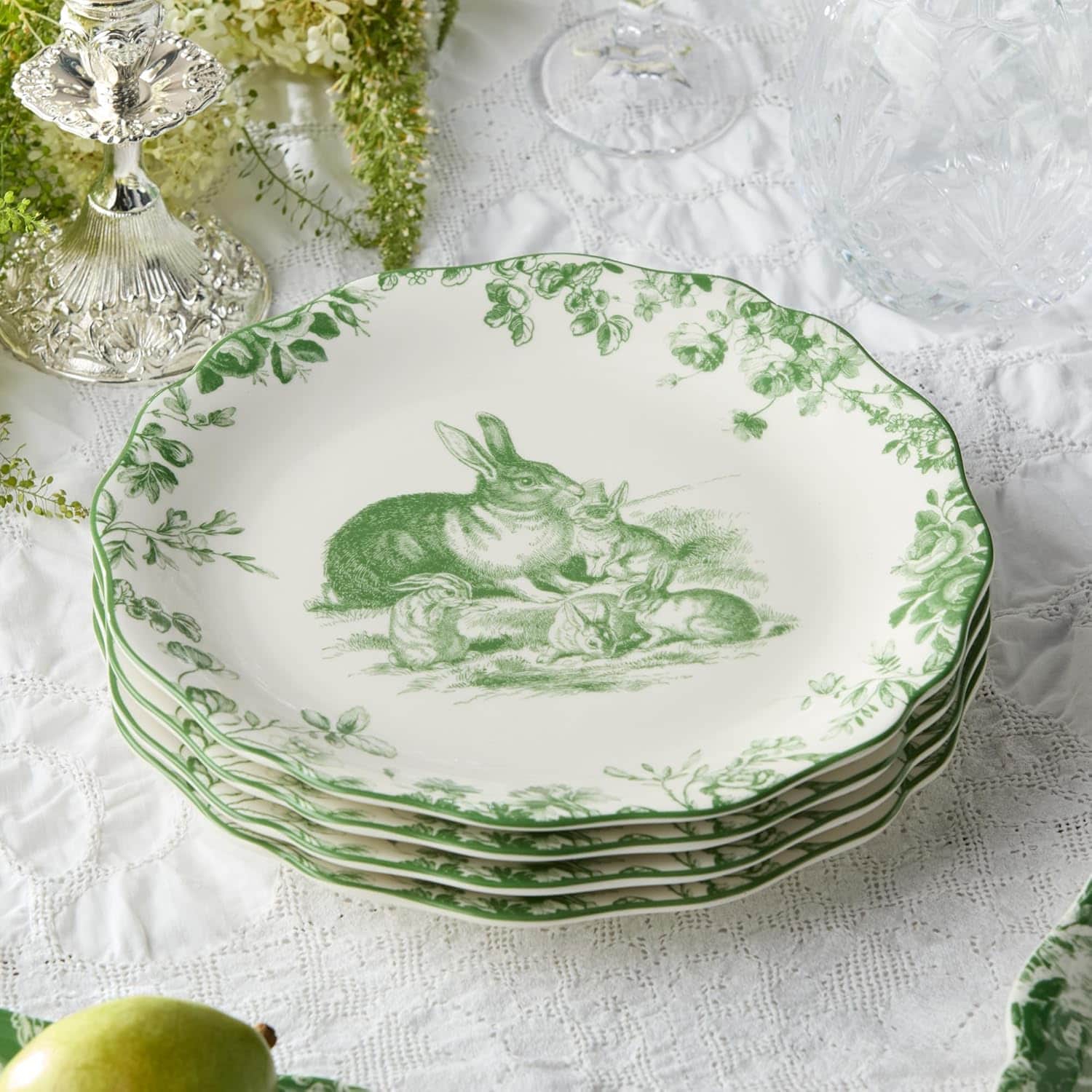 A stack of decorative plates with green illustrations of rabbits, evoking the freshness of first greens, set on a white lace tablecloth with glassware and floral accents nearby.