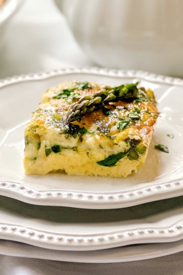 A square slice of vegetable frittata, garnished with asparagus and herbs, is served on a white plate—an ideal choice for spring recipes.