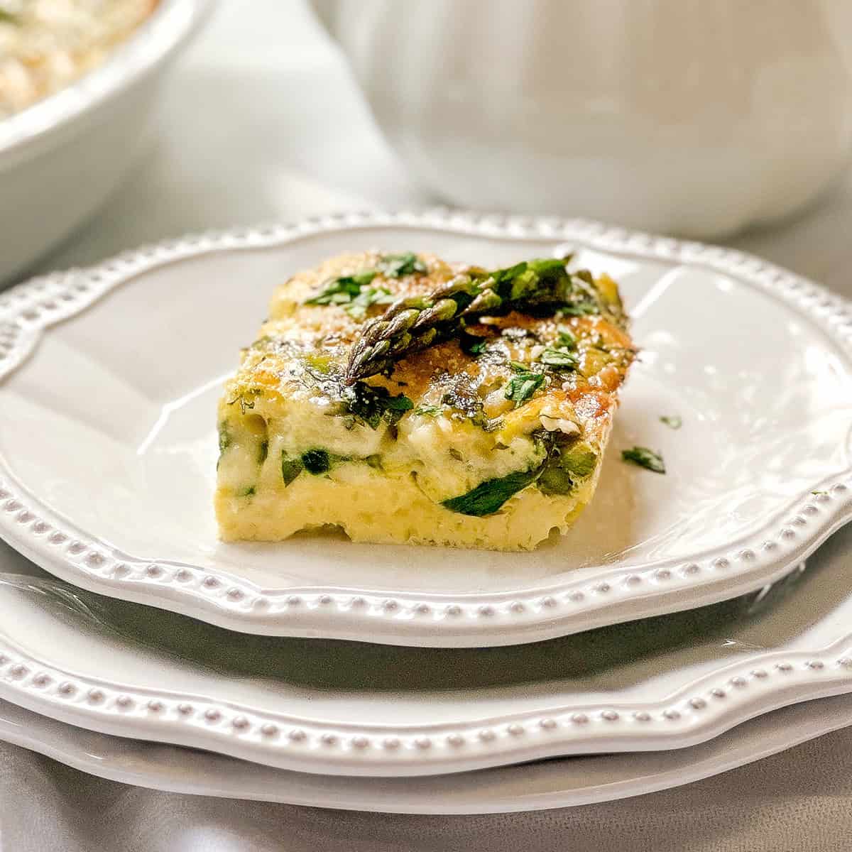 A square slice of vegetable frittata, garnished with asparagus and herbs, is served on a white plate-an ideal choice for spring recipes.