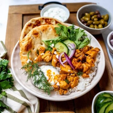 A bowl with naan, quick chicken shawarma, rice, fresh greens, cucumber, red onion, dill, yogurt sauce, and a side of green olives on a wooden tray.