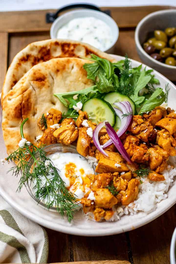 A bowl with naan, quick chicken shawarma, rice, fresh greens, cucumber, red onion, dill, yogurt sauce, and a side of green olives on a wooden tray.