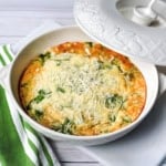 A baked spinach and cheese frittata sits in a white casserole dish, topped with shredded cheese, next to a green and white striped napkin—perfect for those who love quick and easy recipes.
