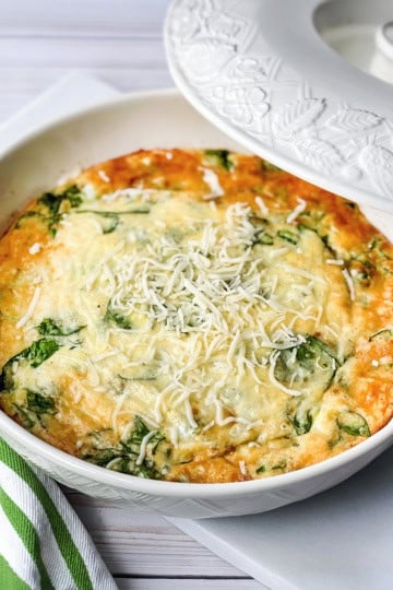 A baked spinach and cheese frittata sits in a white casserole dish, topped with shredded cheese, next to a green and white striped napkin-perfect for those who love quick and easy recipes.
