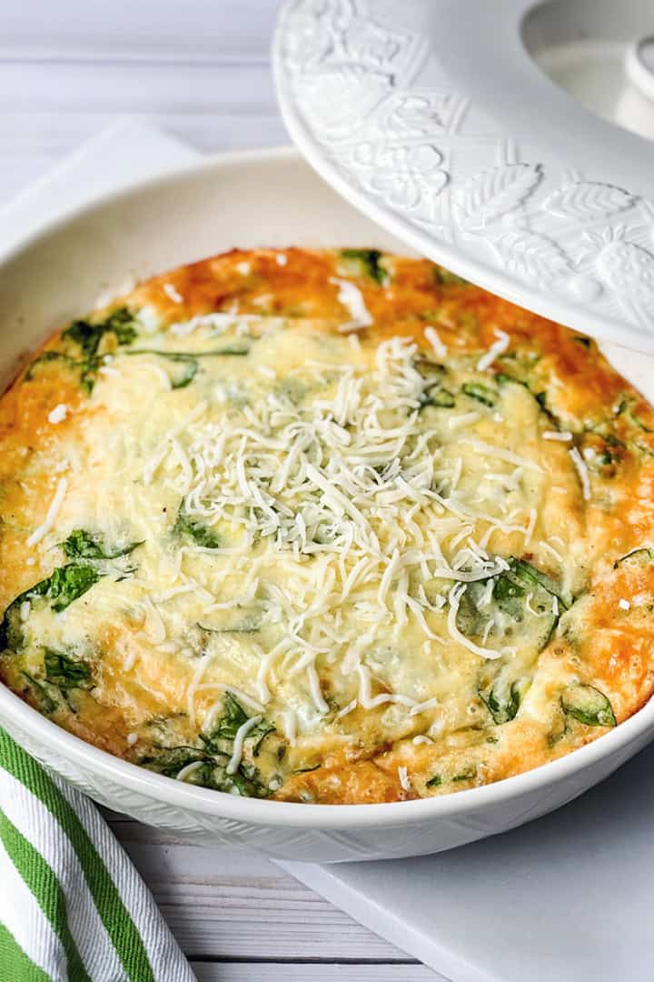 A baked spinach and cheese frittata sits in a white casserole dish, topped with shredded cheese, next to a green and white striped napkin—perfect for those who love quick and easy recipes.