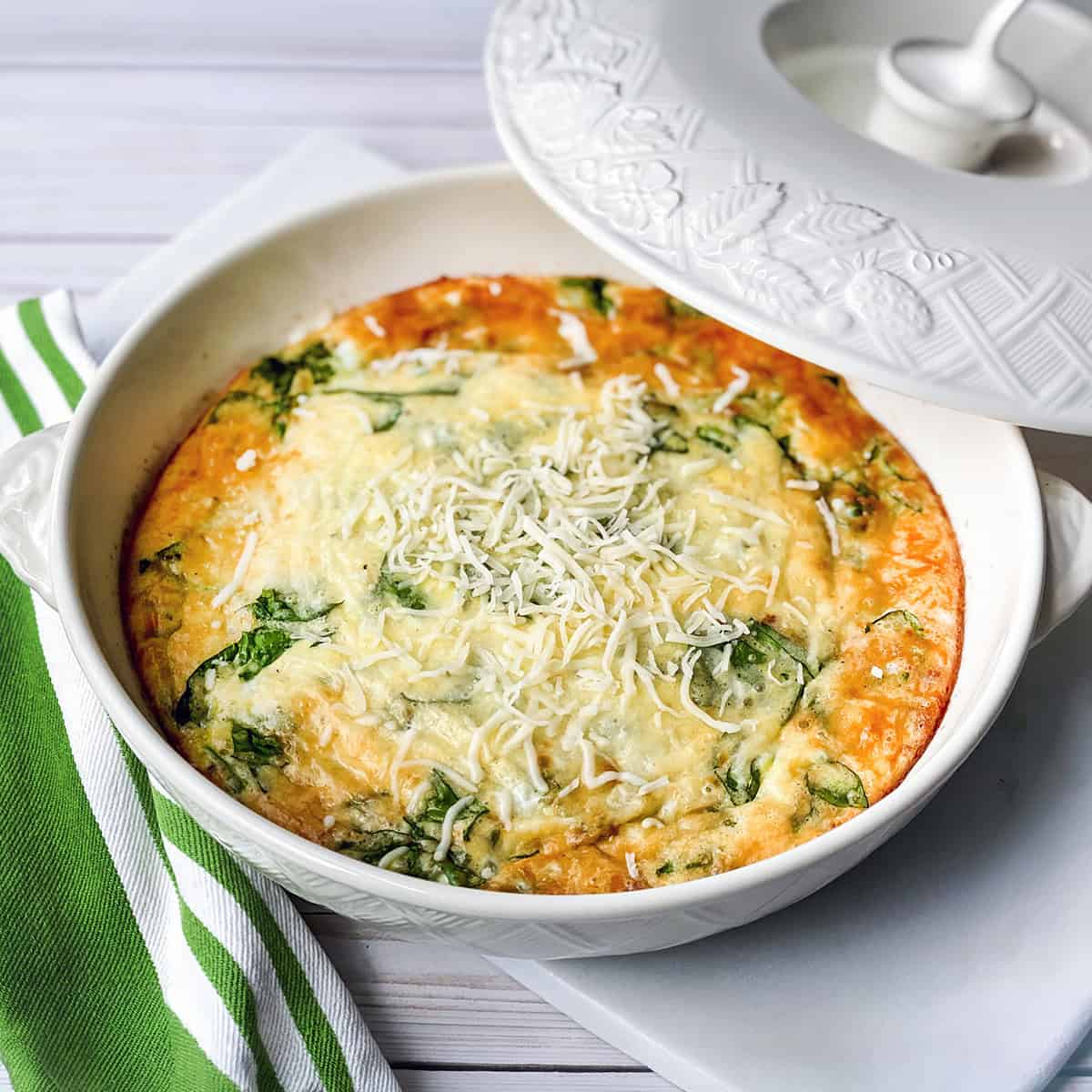 A baked spinach and cheese frittata sits in a white casserole dish, topped with shredded cheese, next to a green and white striped napkin—perfect for those who love quick and easy recipes.