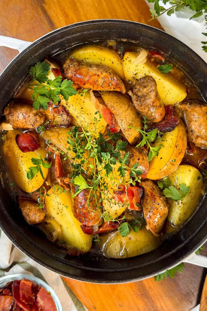 A black pot filled with roasted sausages, potato wedges, tomatoes, onions, and fresh herbs-reminiscent of a hearty Dublin Coddle-rests on a wooden surface beside a striped towel and a bowl of cooked bacon.