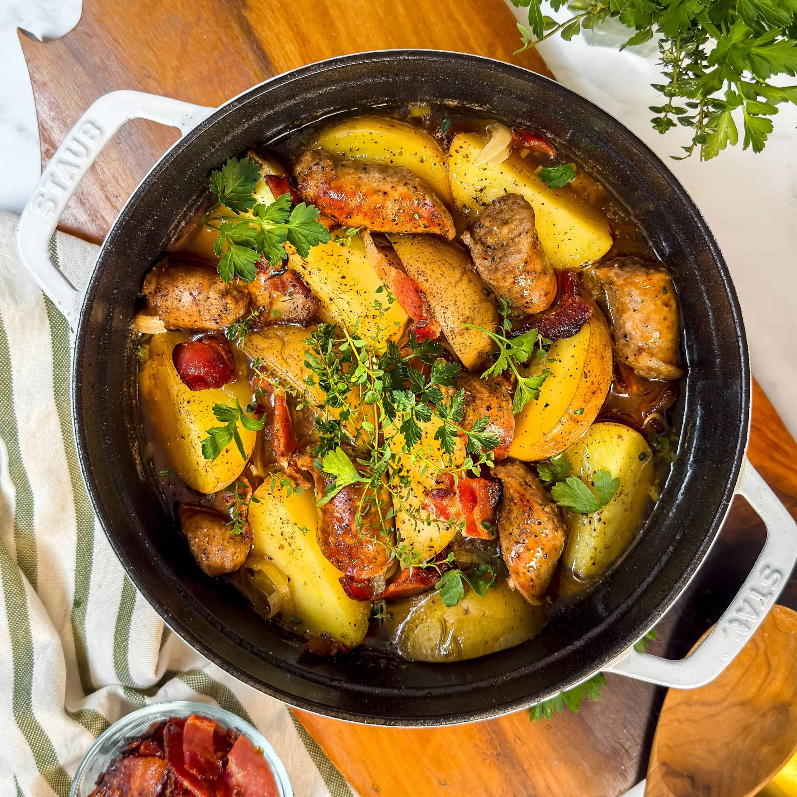 A black pot filled with roasted sausages, potato wedges, tomatoes, onions, and fresh herbs—reminiscent of a hearty Dublin Coddle—rests on a wooden surface beside a striped towel and a bowl of cooked bacon.