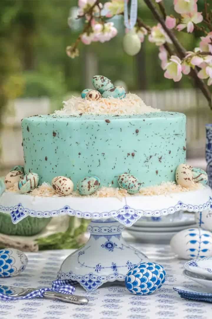 A light blue frosted cake with decorative speckled eggs sits on a patterned cake stand, surrounded by blue and white china and Easter-themed decor-perfect for an elegant Easter tea party on an outdoor table.