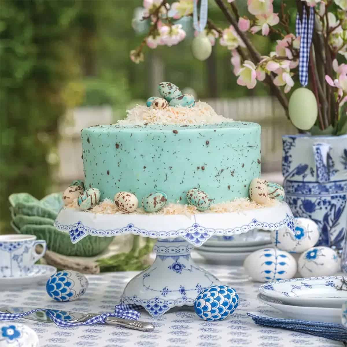 A light blue frosted cake with decorative speckled eggs sits on a patterned cake stand, surrounded by blue and white china and Easter-themed decor—perfect for an elegant Easter tea party on an outdoor table.