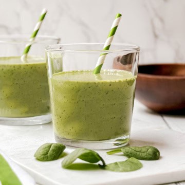 A glass of green smoothie, perfect for your first greens boost, with a striped paper straw sits among fresh spinach leaves on a white surface. Another glass and a wooden bowl appear in the background.