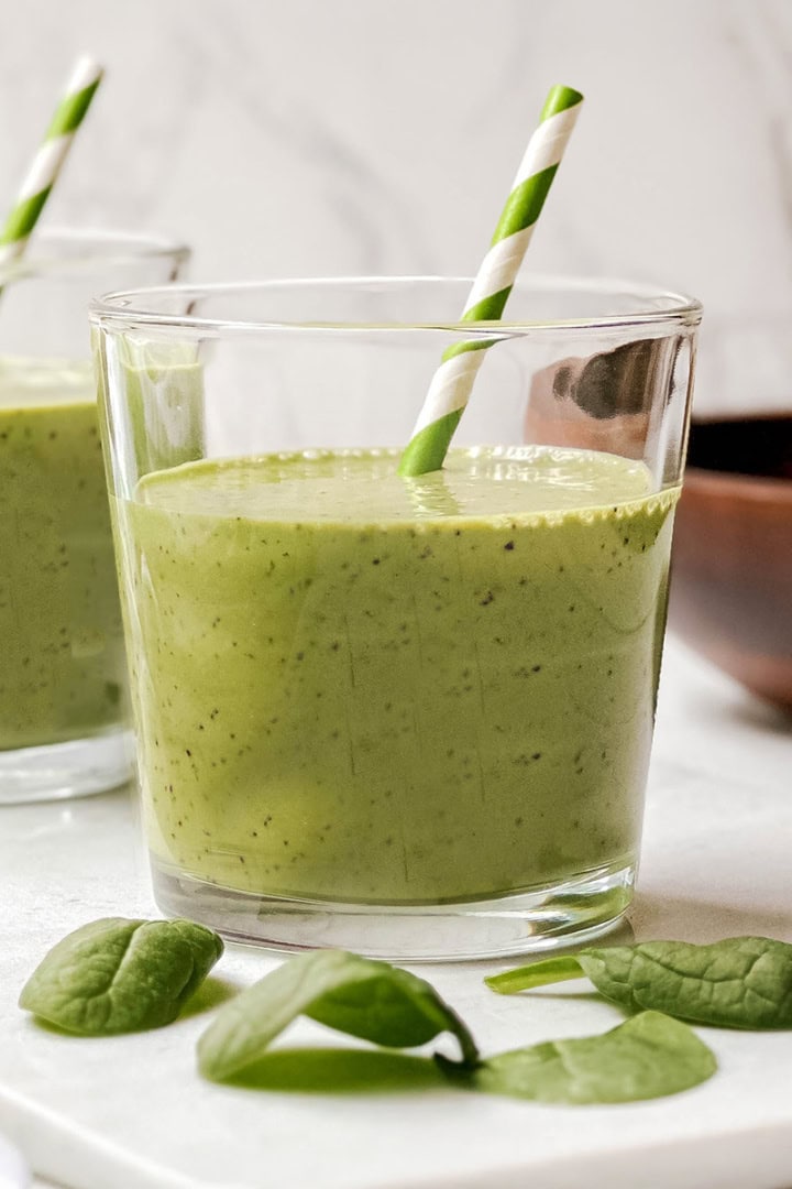 A glass of green smoothie, perfect for your first greens boost, with a striped paper straw sits among fresh spinach leaves on a white surface. Another glass and a wooden bowl appear in the background.