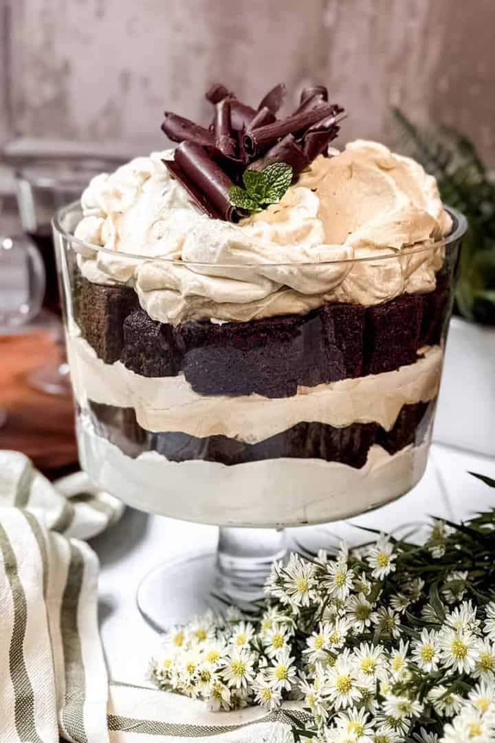 A glass trifle dish filled with layered chocolate cake and whipped cream, topped with chocolate curls and a mint sprig, makes a festive choice for St Patrick's Day Desserts, surrounded by flowers and a striped cloth.