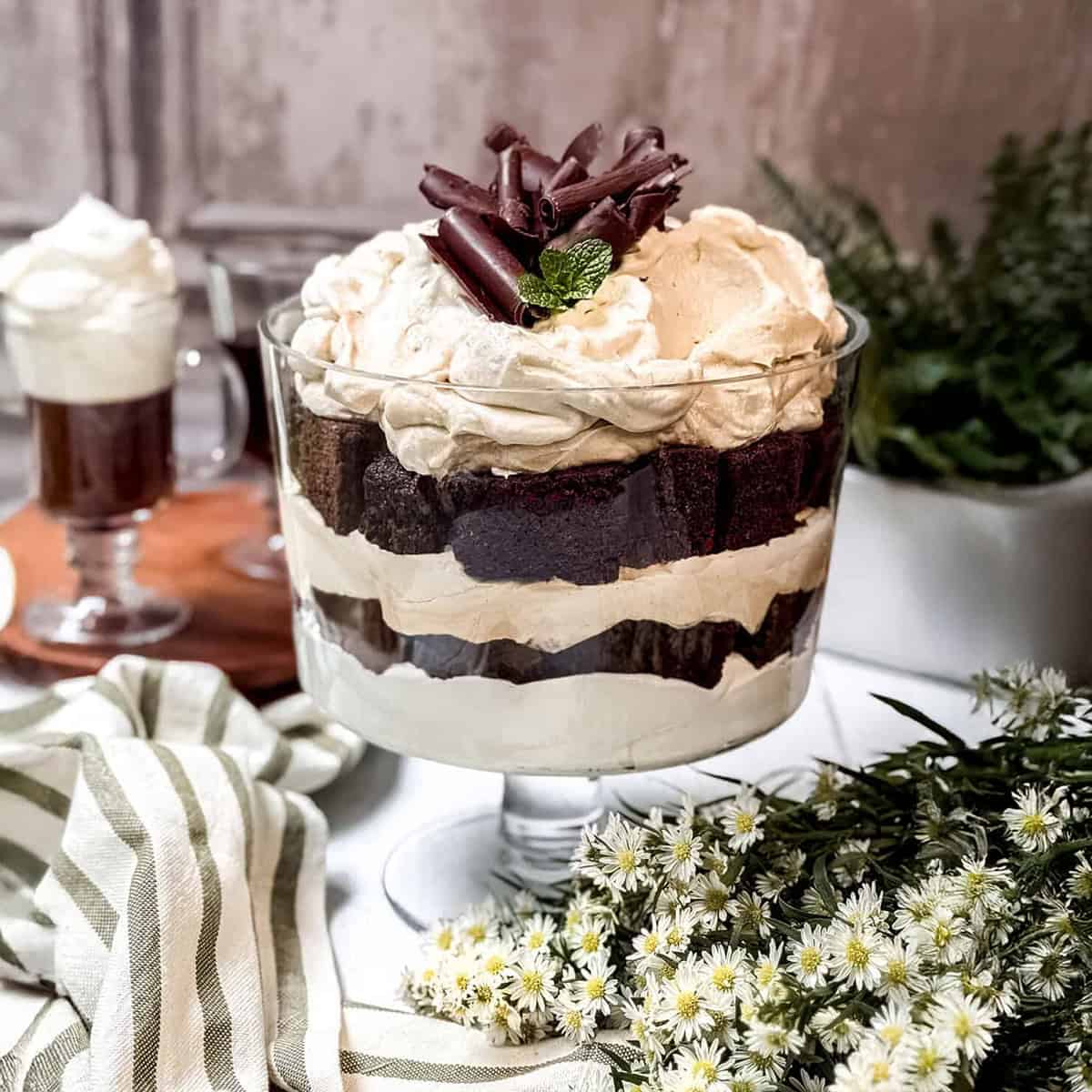 A glass trifle dish filled with layered chocolate cake and whipped cream, topped with chocolate curls and a mint sprig, makes a festive choice for St Patrick's Day Desserts, surrounded by flowers and a striped cloth.
