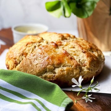 A round loaf of golden-brown bread sits on parchment paper beside a green-striped cloth and a small white flower, capturing the warmth as the season begins, with a cup and potted plant in the background.