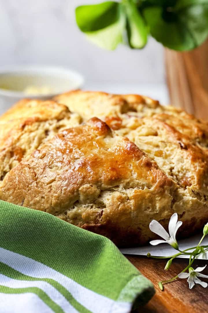 A round loaf of golden-brown bread sits on parchment paper beside a green-striped cloth and a small white flower, capturing the warmth as the season begins, with a cup and potted plant in the background.