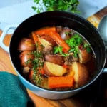A pot of Irish Stew with beef, red potatoes, carrots, herbs, and broth sits on a wooden surface, garnished with parsley.