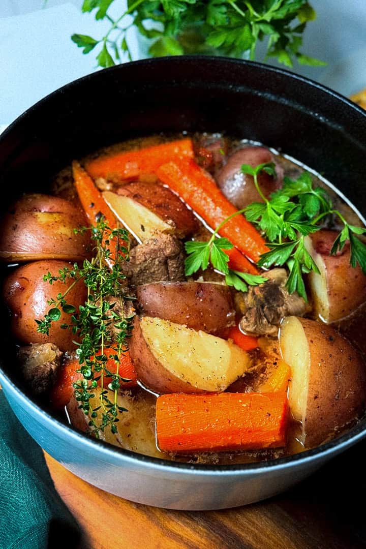 A pot of Irish Stew with beef, red potatoes, carrots, herbs, and broth sits on a wooden surface, garnished with parsley.