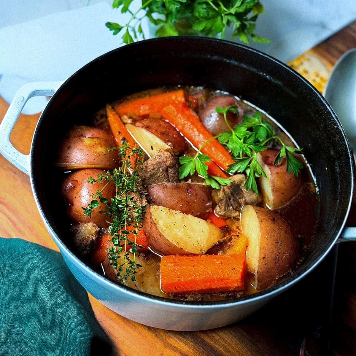 A pot of Irish Stew with beef, red potatoes, carrots, herbs, and broth sits on a wooden surface, garnished with parsley.
