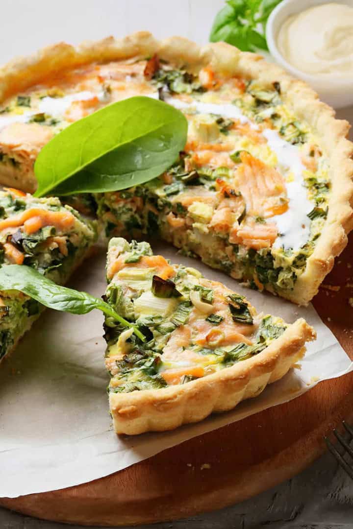 A spinach and salmon quiche on a wooden board, with one slice partially removed. Perfect for March light and fresh dinners, it's served with a small bowl of sauce and some greens in the background.