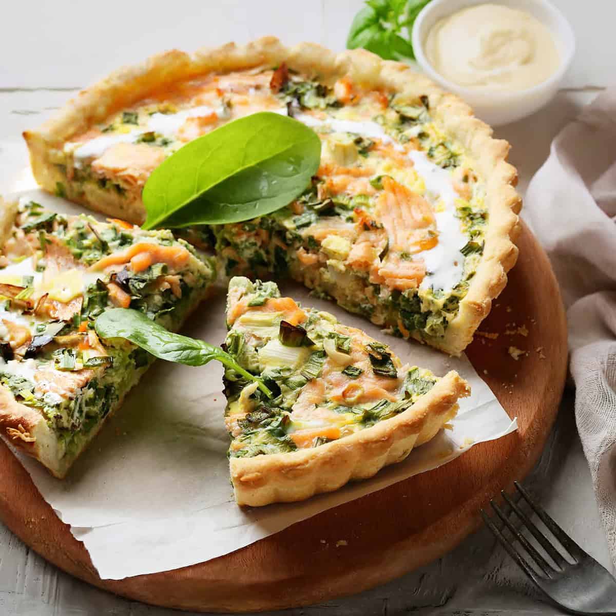 A spinach and salmon quiche on a wooden board, with one slice partially removed. Perfect for March light and fresh dinners, it's served with a small bowl of sauce and some greens in the background.