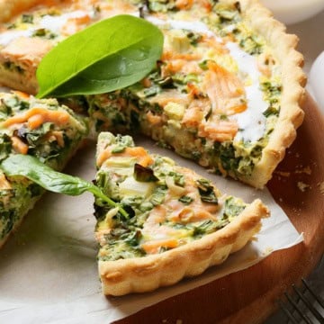 A salmon and dill quiche with a slice cut out, garnished with fresh spinach, sits on a wooden board lined with parchment paper.