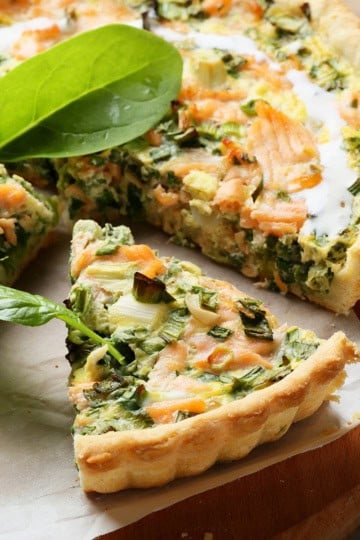 A salmon and dill quiche with a slice cut out, garnished with fresh spinach, sits on a wooden board lined with parchment paper.