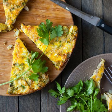Sliced vegetable frittata on a wooden board with fresh cilantro and first greens garnish, a knife, and additional slices on a plate.