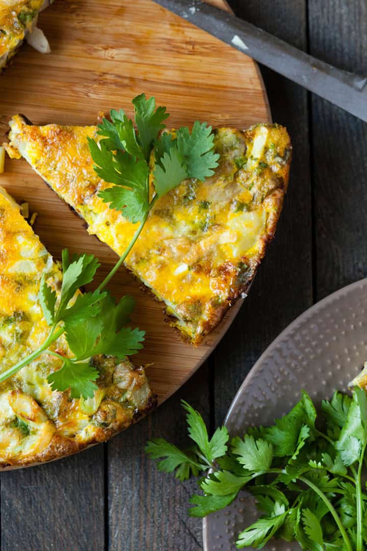 Sliced vegetable frittata on a wooden board with fresh cilantro and first greens garnish, a knife, and additional slices on a plate.